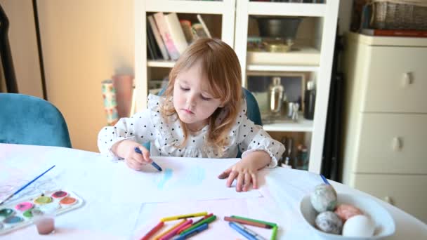 小さな女の子は食堂のテーブルに座って 図面を作成し 創造的であること 彼女は紙の上に青いクレヨンで絵を描いている — ストック動画