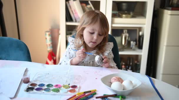 小女孩坐在餐桌旁 画着复活节彩蛋 画着图画 还很有创意 她正在把一只蛋漆成黑色 — 图库视频影像