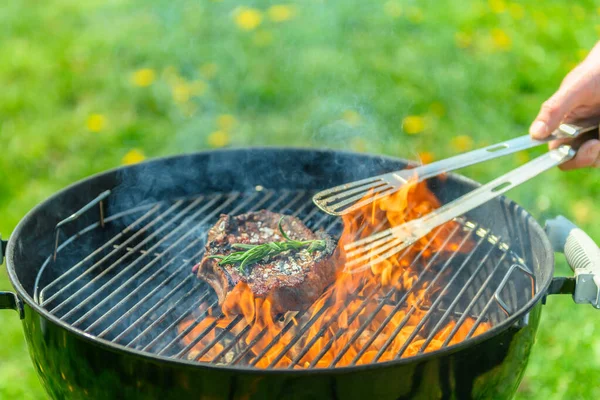 Delicious, juicy sirloin roasting on a traditional black grill, with a lot of fire in the grill. There is a lush green lawn with dandelions in the background. A hand reaches in to grab the steak with a grill tong. There is rosemary on the steak