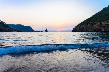 İspanya, İbiza 'nın kuzeybatısındaki Benirras sahilinde güzel bir günbatımı. Sahilde küçük dalgalar dolaşıyor. Arka planda, Tanrı 'nın Parmağı olarak da bilinen Kaptan Bernat' ın kaya oluşumu görülebilir.