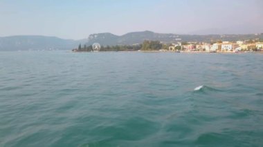 Garda Gölü 'ndeki Bardolino limanından Peschiera del Garda' ya giden turist feribotu. Arka planda büyük, beyaz dönme dolap var. Güneşli bir yaz günü. Yavaş çekim 4K