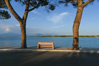 Peschiera del Garda 'dan görülen Garda Gölü' nün muhteşem manzarası. Manzaranın tadını çıkarabileceğin iki büyük ağacın arasında bir park bankı var. Çok güzel bir yaz akşamı.