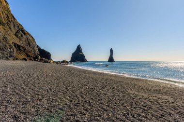 Reynisdrangar kayalıkları İzlanda 'daki Reynisfjara siyah kumlu plajında. Uçurumların etrafında uçan bir sürü kuş, onların üreme alanı. Güneşli bir bahar günü ve Atlantik Okyanusu 'ndan gelen hafif dalgalar sahile vuruyor.