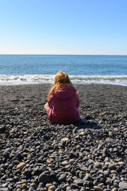 İzlanda 'da Reynisfjara' daki güzel okyanus manzarasını izleyen küçük, tatlı bir kız. O, sırtı kameraya dönük olarak kum ve çakıl üzerinde oturuyor, Atlantik Okyanusu 'nu izliyor. Güneşli bir bahar günü.