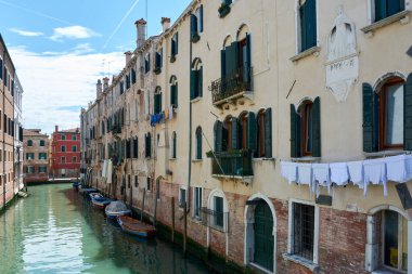 Picturesque facades of old buildings with hanging clothes and boats parked on canal in Venice, Italy, under blue sky with clouds on spring day. Beautiful view in famous Italian landmark.