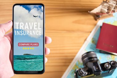 Top view of male hand holding mobile phone with travel insurance website on the screen. Travel items in the background with camera, passport and map. Travel insurance website or app concept. Copy space. clipart