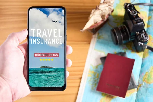 Top view of male hand holding mobile phone with travel insurance website on the screen. Travel items on wooden table in the background with camera, passport and map. Travel insurance website or app concept.