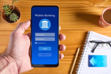 Ekranda online bankacılık uygulaması arayüzü olan cep telefonu, kullanıcı adı ve şifre kutularıyla giriş sayfası, kredi kartı ve aksesuarları arka planda bulanık olan ahşap masa üzerinde bulunan erkek el telefonu.