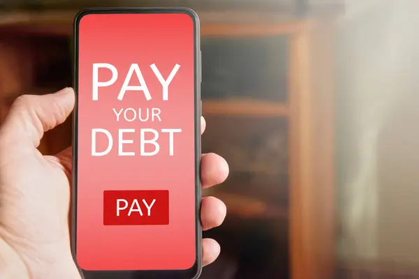 Man holding mobile phone with pay your debt text and pay button on red screen. Online debt payment on smartphone. Financial problem.