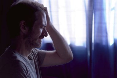 Üzgün, genç, sakallı bir adam gözleri kapalı ve eli kafasında ağlıyor. Pencereli loş ışık. Depresyon ve akıl sağlığı kavramı.