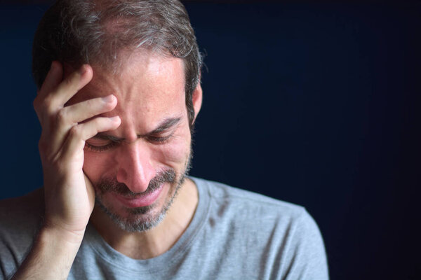 Close up of young caucasian 30 year old man with short beard and blue eyes crying while holding his head. Concept of depression, loss and grief. Emotional trauma.