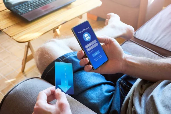 Close up of man on the couch managing finances with a mobile phone, navigating online banking with a credit card in hand. Modern, convenient and secure financial transactions at home.