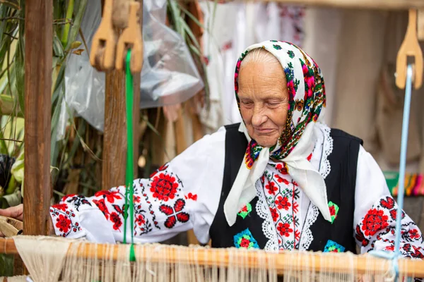 An old Belarusian or Ukrainian woman in an embroidered shirt at a vintage loom. Slavic elderly woman in national ethnic clothes.