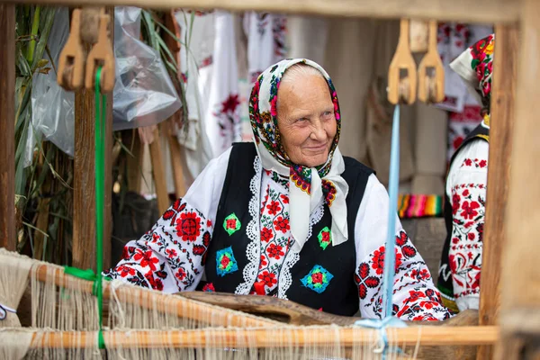 An old Belarusian or Ukrainian woman in an embroidered shirt at a vintage loom. Slavic elderly woman in national ethnic clothes.