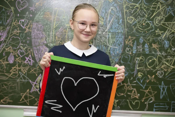 A schoolgirl of middle school age in glasses holds a slate board on which a heart is drawn.