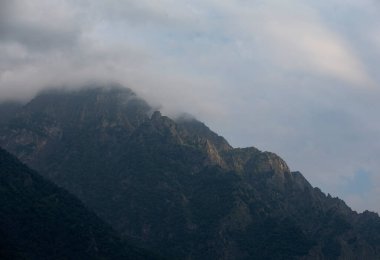 Karanlık atmosferik gerçeküstü manzara. Koyu kayalık bir dağ zirvesi. Alçak bulutlar ve gri bulutlu bir gökyüzü. Yüksek bir tepede gri bir alçak bulut. Alçak bulutlarda yüksek siyah kaya. Gerçeküstü kasvetli dağlar.