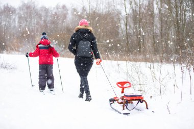 Oğlu kayak yapıyor, annesi de kızağı arkaya itiyor. Ormanda kış yürüyüşü.