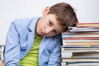 Yorgun bir okul çocuğu kitap yığınlarının arasında oturur. Kitapları olan üzgün çocuk..
