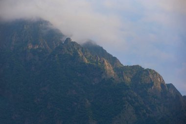 Karanlık atmosferik gerçeküstü manzara. Koyu kayalık bir dağ zirvesi. Alçak bulutlar ve gri bulutlu bir gökyüzü. Yüksek bir tepede gri bir alçak bulut. Alçak bulutlarda yüksek siyah kaya. Gerçeküstü kasvetli dağlar.