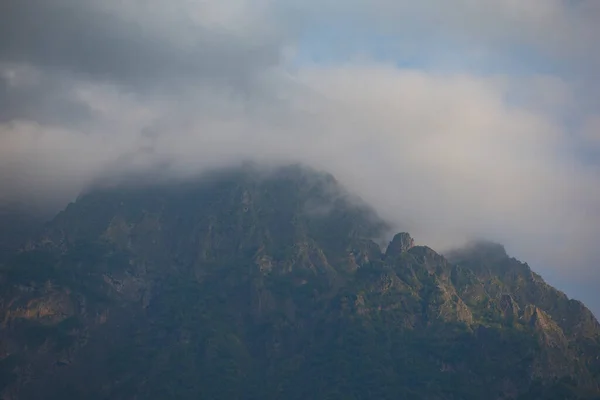 Stock image Dark atmospheric surreal landscape with a dark rocky mountain peak in low clouds in a gray cloudy sky. A gray low cloud on a high peak. High black rock in low clouds. Surreal gloomy mountains.