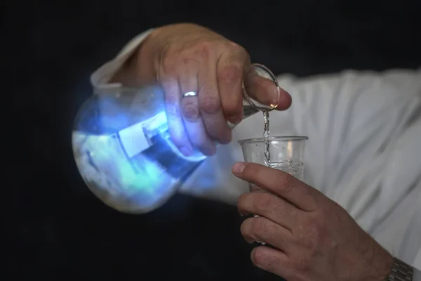 青色の溶液と実験室のアシスタントの手に神秘的な輝きを持つ化学フラスコ 経験と実験 — ストック写真
