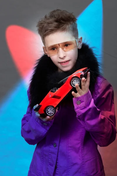 おしゃれな男の子が多色のスタジオを背景におもちゃの車を手に持っています 高価な車を夢見ている未来的なメガネの美しい子供 子供の夢 金持ちの子 — ストック写真