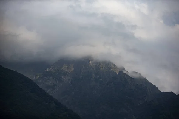 stock image Dark atmospheric surreal landscape with a dark rocky mountain peak in low clouds in a gray cloudy sky. A gray low cloud on a high peak. High black rock in low clouds. Surreal gloomy mountains.