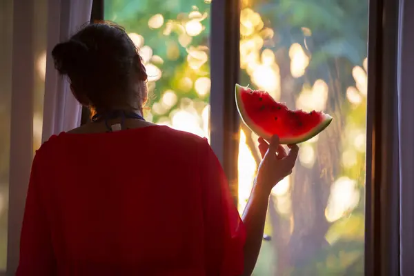 Silhouette of a woman at the window with a watermelon in the rays of the setting sun.
