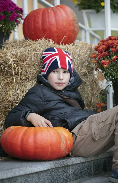 A boy in a hat with the English flag sits with large pumpkins.