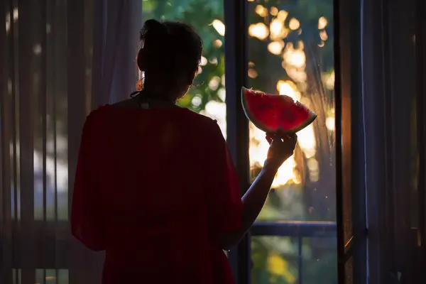 Silhouette of a woman at the window with a watermelon in the rays of the setting sun.