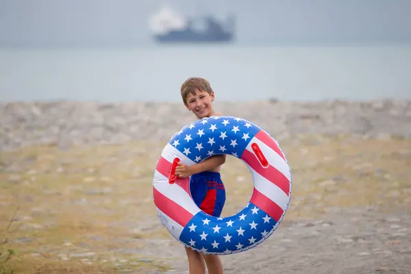 A happy boy wearing swimming goggles and an inflatable life buoy with the American flag painted on it.