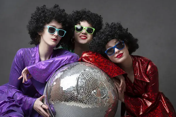 A group of disco girls in wigs with a disco ball and colorful costumes pose against a gray background. Disco party.