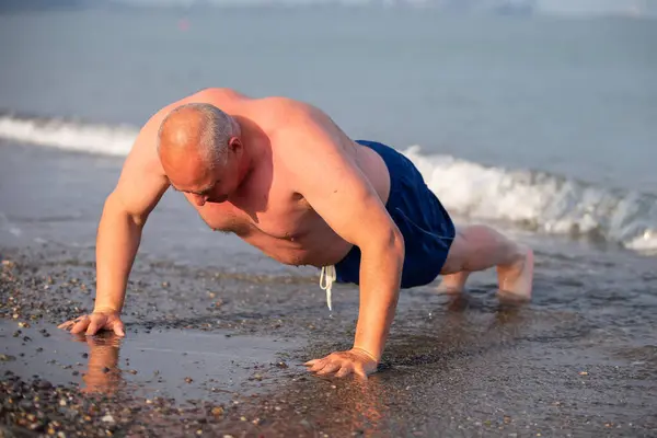 An elderly man of athletic build plays sports on the shore. Mature man doing push-ups outdoors.