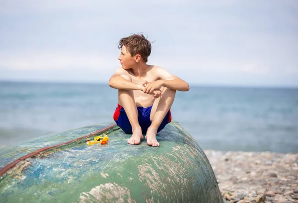 A boy on the beach sits on an old boat washed ashore by a storm.