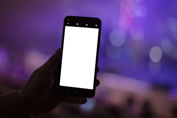 A hand holds a smartphone with a blank screen to copy your text or information content on a dark concert bokeh background.