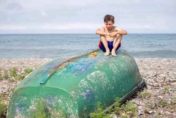 A boy on the beach sits on an old boat washed ashore by a storm.