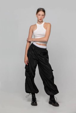 Açık tişörtlü ve siyah geniş pantolonlu tam boy on yedi yaşında bir kız. Kameraya poz veren manken kız.