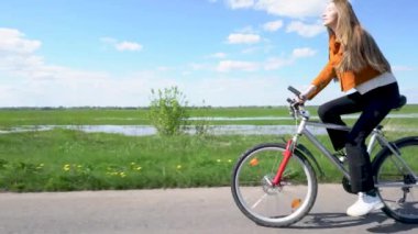 Bir kız güzel bir tarla manzaralı bir yol boyunca bisiklet sürüyor. Saçları uçuyor ve tekerlek de dönüyor..