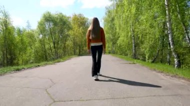 Uzun saçlı genç bir sarışın bir huş ağacının yanından asfalt yol boyunca sırtı kameraya dönük yürüyor..