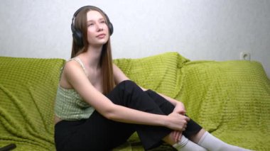 Kanepede oturan genç kız kulaklıkla müzik dinliyor, başını sallıyor ve melodinin tadını çıkarıyor..