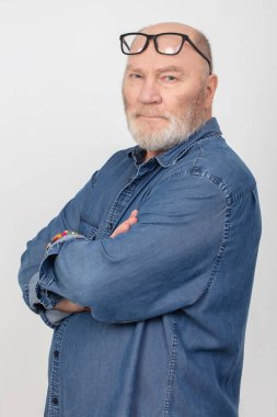 Gri arka planda izole edilmiş kot gömlek giyen, gözlüklü, beyaz saçlı çekici bir adamın portresi. Avrupalı sakallı büyükbaba..