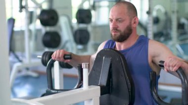 Bir adam ağırlık makinesiyle çalışıyor. Mavi bir atlet giyiyor ve sakalı var.