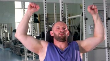 Bir adam ağırlık makinesiyle çalışıyor. Mavi bir atlet giyiyor ve sakalı var.