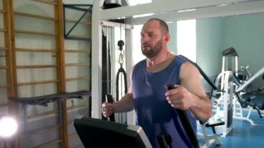 Spor salonunda eliptik bir antrenörde erkek bir sporcu. Kolları hareket ettiriyor. Spor salonu çeşitli egzersiz makineleriyle dolu..