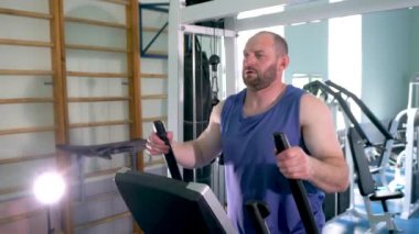 Spor salonunda eliptik bir antrenörde erkek bir sporcu. Kolları hareket ettiriyor. Spor salonu çeşitli egzersiz makineleriyle dolu..