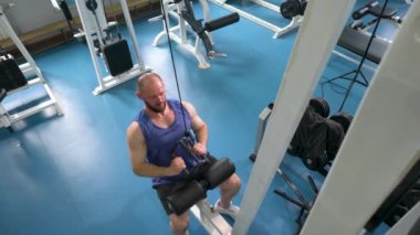 Spor salonunda ağırlık makinesi kullanan bir adam. Spor salonu temiz ve temiz, mavi ve beyaz renk düzeniyle. Adam antrenmanına odaklanmış..