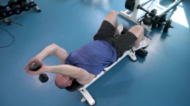 Bir adam spor salonunda ağırlık kaldırıyor. Mavi bir atlet ve şort giyiyor. Sağ elinde bir dambıl tutuyor.