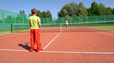 Bir bayan koç tenis kortunda bir erkek sporcuyla konuşur. Ellerinde raket ve toplar var.