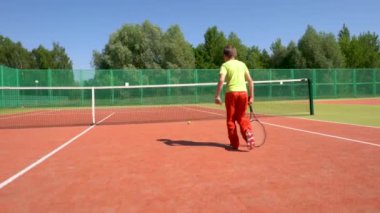 Genç bir çocuk tenis kortunun etrafında raketle koşup topa vuruyor..
