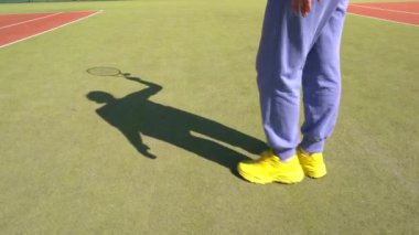 Haritada tanınmayan bir kadın tenis kortunu vuruyor. Eşofman altı ve sarı spor ayakkabı giyiyor..